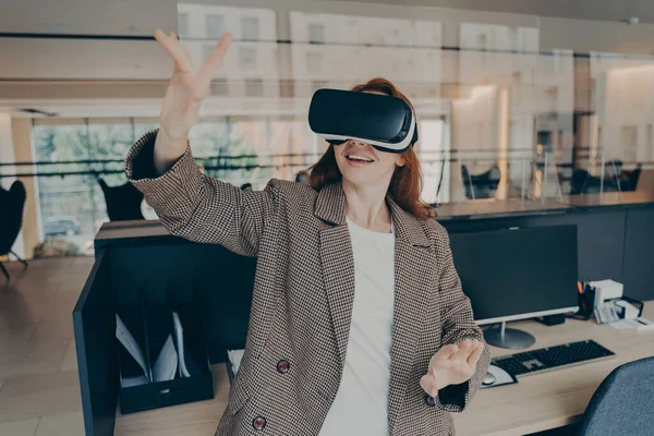 Female office worker using virtual reality to visualize and browse project files, using her hands to control objects in digital world, excited business woman wearing vr headset gesturing in office
