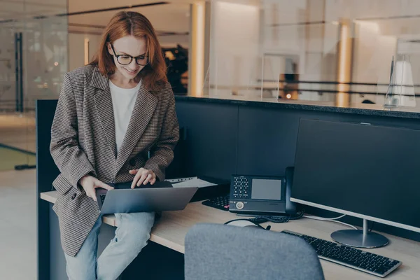 ノートパソコンを使用して作業机に座って インターネットを閲覧し 現代のオフィスのインテリアで働くコンピュータ 赤毛の女性を使用して研究を行う忙しい女性は 現代の技術を使用して — ストック写真
