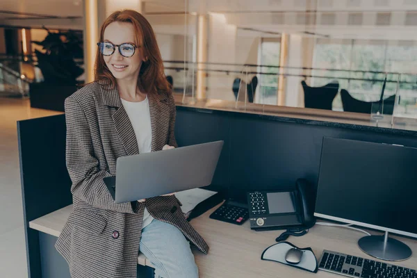 現代のコワーキングスペースで働きながら ノートパソコンを持ちながら机の上に座ってパソコンを見回したり Webをサーフィンしたりする眼鏡をかけた赤髪の女性Olの笑顔 — ストック写真