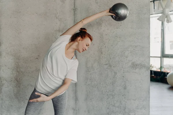Active slim woman doing sport exercises during barre workout, arching body on side and raising hand with small fitball over head against gray wall in fitness studio, being in good physical shape
