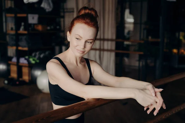 stock image Young beautiful red haired woman with stylish bun leaning on ballet barre in fitness studio with blurred background while smiling and looking into distance, ballerina during stretching workout