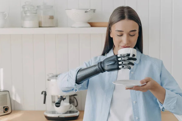 Routine of handicapped person and life quality. Pretty disabled girl is holding cup of tea with cyber hand and drinking. Concept of grasp sensors in modern robotic limb. Woman at domestic kitchen.