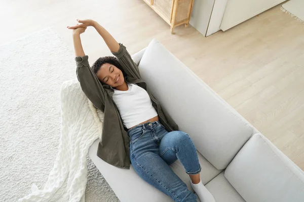 stock image Calm african american young woman lying, stretching body, relaxing on couch in living room breathe fresh air. Happy relaxed girl tenant resting on comfortable sofa at home. Stress relief, wellness