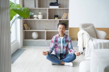 Afrikalı Amerikalı genç bir kadın lotus poz verip meditasyon yapıyor. Genç afro kız evde yoga yapıyor ve nilüfer çiçeği pozisyonu alıyor. Ruhani uyum ve denge. Stres atma..