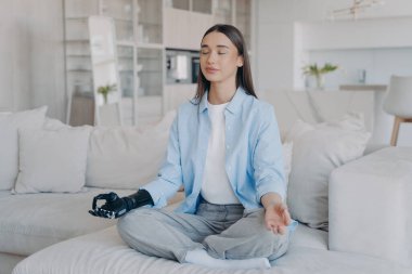 Tranquil disabled woman is sitting in lotus pose on couch with her eyes closed. Attractive caucasian girl is amputee. Lady has myoelectric cyber prosthesis. Modern artificial limb with sensor control.