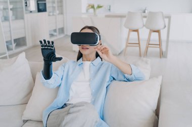 Disabled girl with cyber arm in vr glasses. Handicapped person gets rehabilitation at home. Young attractive european woman is sitting in living room on couch. Healthcare innovation technology.