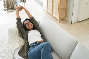 Lazy weekend morning of teenager. Carefree african american girl is relaxing on couch and pillow at her apartment. Young afro woman is stretching and smiling. Leisure and rest concept.