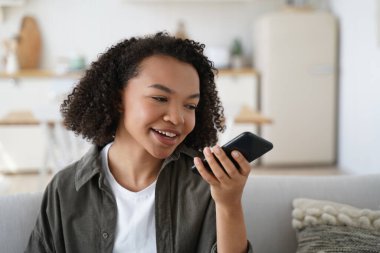 Friendly african american young girl sit on couch at home hold smartphone leave voice message. Modern biracial female chat with friends use loudspeaker virtual assistant search information on internet
