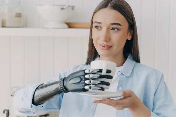 Confident handicapped girl is holding cup of tea with cyber hand. Happy disabled girl at kitchen at home. Pretty woman with makeup is smiling. Concept of grasp sensors in modern electronic prosthesis.