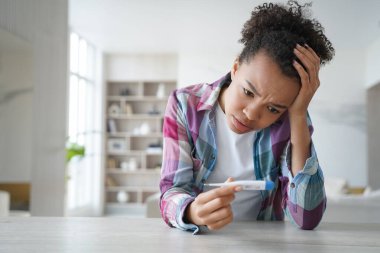Anxious teenage girl gets positive pregnancy test result. Desperate african american young woman is holding stripe test and looking at it. Teenage lifestyle and mistakes. Contraception concept.