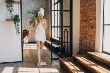 Anxious slim caucasian woman taking step onto scale to measure her weight. Girl wrapped in towel after bathing has time at spa. Young woman checking weight after shower. Fitness and wellness concept.