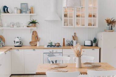 İskandinav mutfak tasarımı. Modern apartman dekorasyonu. Ev mutfağı ya da yemek odası. Beyaz, lüks iç mekan gün ışığında aydınlanıyor. Mutfak aletleri, mutfak gereçleri, soba, masa ve buzdolabı..