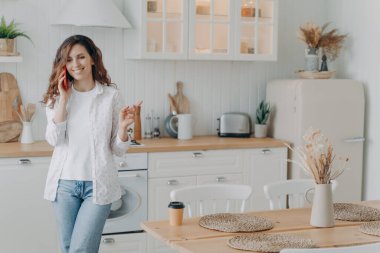 Young european woman talking on smartphone in her stylish kitchen at home. Happy attractive housewife has leisure. Modern white scandinavian interior. Stove, worktop and cuisine.