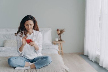 Genç ve güzel bir bayan evde telefonla konuşuyor. Sıradan giyinen mutlu Avrupalı kız mesajlaşıyor ve mesajlar alıyor. Boş zaman ve iletişim konsepti. Evcil kız iyi vakit geçiriyor..