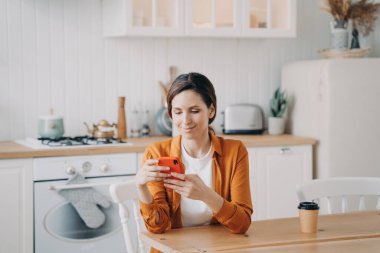 Happy european girl is sitting at the table at kitchen and texting on phone. Young woman in orange shirt is remote worker. Lady has coffee and browsing through internet. Workplace at home.