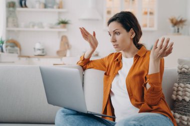 Puzzled young woman has online conference at home. European lady with laptop is talking at internet meeting. Confused girl is telling a problem. Businesswoman is working while quarantine.