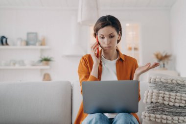 Young caucasian woman has laptop problem. Girl has consultation on phone. Lady is talking on telephone and telling some problem. Puzzled woman is sitting on sofa at home holding computer.