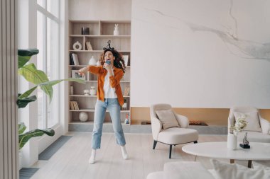 Heyecanlı kız oturma odasında eğleniyor. Mutlu Avrupalı kadın mikrofondaki gibi evde telefonla şarkı söylüyor. Favori şarkı söylüyor. Hafta sonu sabahı tek başına dans etmek.