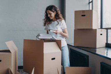 Curly young woman, student is housing in residential room. Happy european woman unpacking boxes with books. Girl in white t-shirt indoors. Relocation and independence concept.