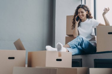 Happy hispanic woman is sitting on windowsill of new house. Girl is unpacking boxes and talking on phone to friends. Relocation and communication concept.