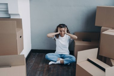 Exhausted young woman amond packed boxes. Anxious girl is holding her head with hands. European woman getting trouble. Stress, headache and depression from moving concept.