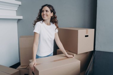 Hopeful lady is holding cardboard box and moving to modern apartment. Happy european woman purchasing real estate and looking at interior. Improvement, changes and new life concept.