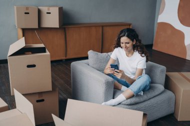 Young spanish woman posting news. Happy apartment buyer unpacking boxes. Girl is moving to new home. Relaxed young lady clicks mobile phone. Independence and achievement concept.