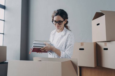 Female student moves to campus room. Young european woman unpacking carton boxes and takes textbooks out. Happy girl in glasses unloading luggage. Relocation and entering to college.