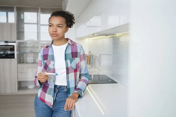 Pensive teenage girl gets positive pregnancy test result. Sad african american young woman is holding stripe test. Teenage lifestyle, unwanted pregnancy and motherhood. Contraception concept.