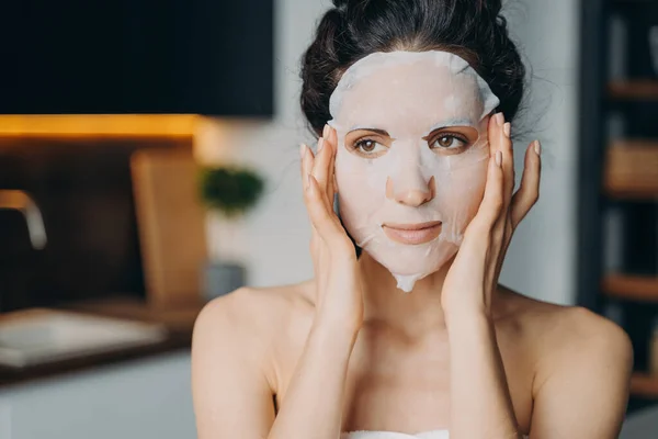 Caucasian girl applies mask sheet after bathing. Vitamin, nourishing and nutrition face mask. Young woman takes shower at home or hotel and doing spa procedures. Skin care and rejuvenation.