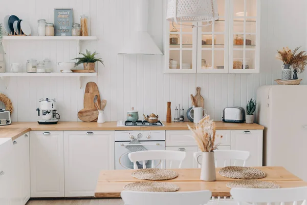 Scandinavian kitchen design. Modern apartment decoration. Domestic kitchen or dining room. White luxurious interior lit by daylight. Kitchen appliance, utensils, stove, table and refrigerator.
