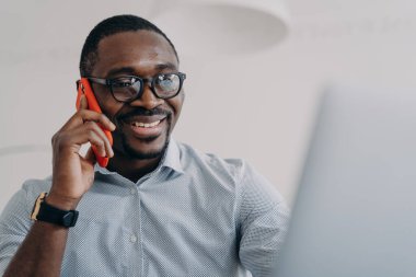 Successful african american businessperson in glasses hold mobile phone makes nice business call sitting in front of laptop. Black male friendly manager consulting a client, talking on smartphone.