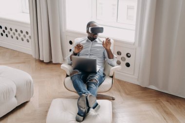 African american man in VR glasses working in augmented reality, sitting in armchair with laptop at home, pointing at virtual reality object, interacting with cyberspace, using modern high-tech device