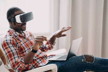 Smiling african american man in VR glasses sits in armchair with laptop, testing virtual reality goggles. Modern black guy viewing purchase in cyberspace. Tech and e-commerce concept.