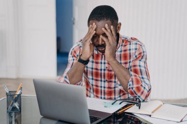 Exhausted african american man suffer headache after working at laptop. Tired frustrated black guy holds head by hands feel migraine pain at workplace. Stress at work, occupational burnout.