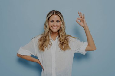 Rahatlamış genç bir bayan, el hareketlerinde ve diğerini kalçasında dinlendirirken, iyi olduğundan emin olarak ve mavi duvarın yanında tek başına dururken her şeyin yolunda gideceğinden emin olarak.