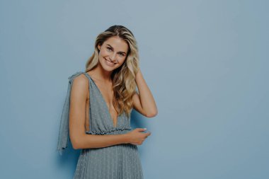 Romantik mavi elbiseli genç sarışın kadın saçlarına hafifçe dokunuyor, eğik kafayla gülümsüyor, mavi arka planda yalnız dururken olumlu duygular ifade ediyor.