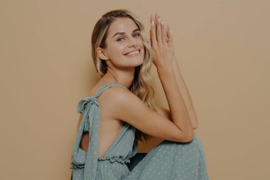 Gece elbisesi içinde zarif, ince sarışın bir kadın katlanır avuç içleriyle sandalyeye oturmuş, hoş bir gülümsemeyle kameraya bakıyor, bej renkli stüdyo arka planında izole edilmiş.