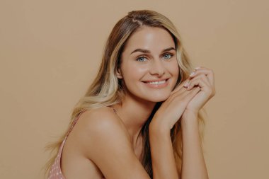 Güzel, romantik sarışın bir kadının stüdyo portresini kapat, kameraya ışıltılı bir gülümsemeyle bak, hassas mavi gözlü, parlak ve saf tenli, yüze yakın elleri olan genç bir kadın.