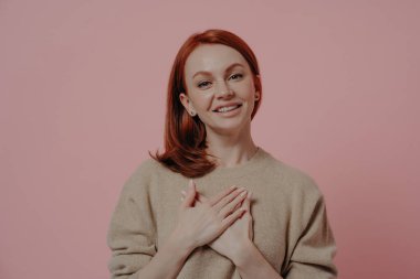 Mutlu, genç, kızıl saçlı bir kadın göğsüne sarılıp teşekkür ediyor. Stüdyoda pembe duvarın önünde dikilirken minnettarlık işareti gösteriyor. Minnettar kızıl kadın kameraya bakıyor.