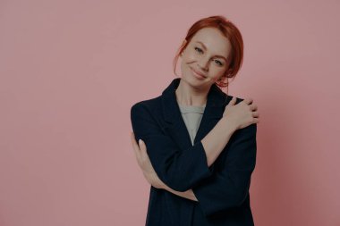 Kendini sevme pratiği. Genç, güzel, kızıl saçlı, gülümseyen, kendine sarılan ve kameraya gülümseyen bir kadın. Pembe pastel arka planda tek başına duruyor. Yazı için fotokopi alanı var.