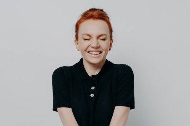 Güzel ve mutlu kızıl saçlı, gözleri kapalı, dişlek gülümsemesi gri duvarda poz veren bir kadın. Neşeli, heyecanlı, pozitif duygular ve mutluluk gösteren, sıradan giyinmiş bir kadın.