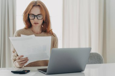 Ciddi kızıl saçlı beyaz kadın, banknotların kağıt belgeler taşıdığını hesaplıyor. Odaklanmış görünüyor. Dizüstü bilgisayar kullanıyor..