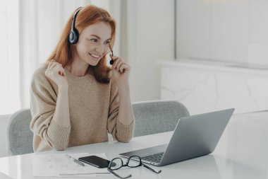 Laptop bilgisayarındaki video görüşmesi sırasında iş arkadaşlarıyla iletişim kuran, kulaklıklı, neşeli, kızıl saçlı bir kadın, evden uzaktan çalışırken online toplantıya katılan bir kadın.