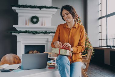 Yeni yıl için dekore edilmiş ofis masasının üstünde oturan neşeli İtalyan iş kadını ve arka planda Xmas ağacı olan neşeli bayan, bayram hediyesi olarak kırmızı kurdeleyi sarıp hediyeye hazırlanıyor.