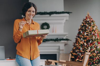 Xmas hediyesi. Kış tatilinde muhteşem bir Noel süslemeli oturma odasında dururken, sıradan hediyeler içinde mutlu Avrupalı bir kadın, paket kutusuna kırmızı kurdele takan bir kadın.