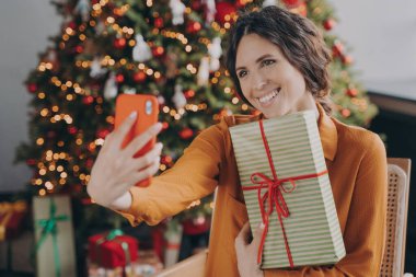 Yeni yıl kutlaması. Neşeli, neşeli İtalyan kadın evde güzel süslenmiş Noel ağacının yanında oturuyor, elinde paketlenmiş Noel hediyesi kutusu ve selfie için poz veriyor, mutlu kadın kış tatilinin tadını çıkarıyor.