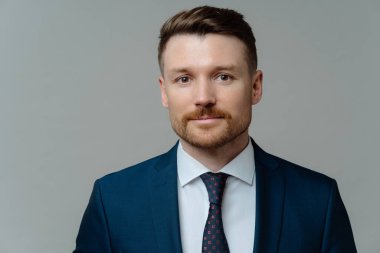 Ciddi özgüvenli iş adamı resmi kıyafetler giyer doğrudan kameraya bakar. Gri stüdyo arka planında izole edilmiş ortaklarla görüşmeye hazır bir görünümü vardır. Motivasyonlu girişimci