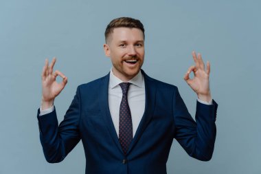 İş başarısı. Mutlu pozitif iş adamı ya da takım elbiseli tatmin olmuş erkek girişimci tamam işareti ya da işaret gösteriyor ve açık mavi arka planda dururken kameraya gülümsüyor.