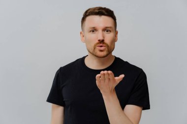 Siyah tişörtlü hoş bir genç adamın portresi hava öpücüğü gönderiyor ve kameraya bakıyor, gri stüdyo arkaplanının üzerinde yalnızken duygularını gösteriyor. Aşk ifadesi kavramı